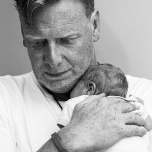 Black-and-white close portrait of a father cradling his sleeping newborn against his shoulder, looking down tenderly.