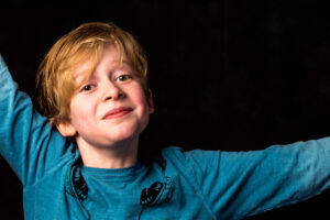 Colour studio portrait of a young boy with strawberry-blonde hair and arms outstretched, wearing a blue T-shirt against a dark background.
