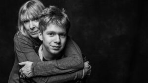 Black-and-white studio portrait of a mother embracing her teenage son from behind, both with relaxed expressions, against a dark background.