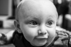 Black-and-white close-up of a baby with wide bright eyes and a soft smile, captured in natural light.