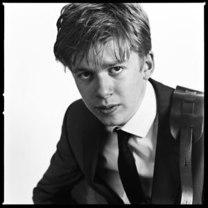 Black-and-white portrait of a young man in suit, tie and waistcoat with a leather strap, direct intense gaze — scanned from medium-format film, vintage character.