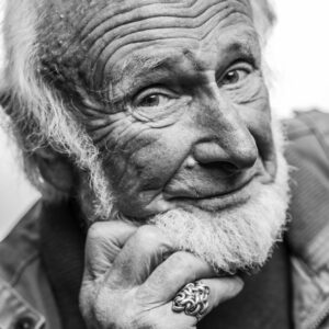 Black-and-white close-up of an elderly man with a long white beard, weathered smiling face and a Celtic-knot silver ring resting against his chin.