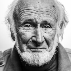 Black-and-white close-up portrait of an elderly man with a long white beard, heavily lined face and pale eyes, contemplative expression.