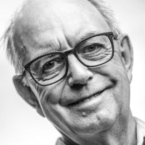 Black-and-white close-up portrait of an older man with grey hair, dark-framed glasses and a broad warm smile, against a bright background.