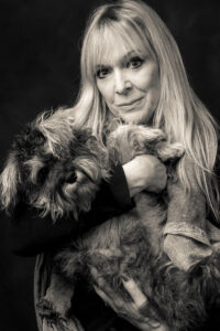 Black-and-white studio portrait of a blonde woman cradling a small grey schnauzer in her arms, both looking towards the camera.