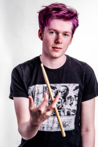 Colour studio portrait of a teenage drummer with vibrant purple hair, holding a drumstick out toward the camera, against a white background.