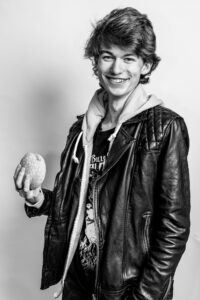 Black-and-white studio portrait of a teenager in a leather jacket and hoody, smiling broadly while holding up a burger.