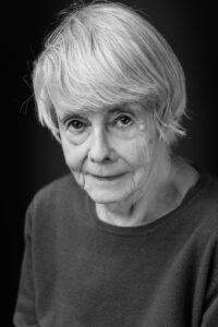 Black-and-white close-up portrait of an older woman with short white hair and a quietly composed expression, against a dark background.