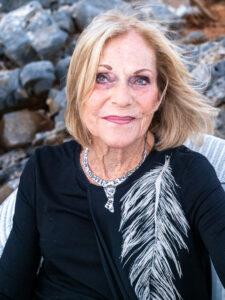 Colour portrait of an older blonde woman wearing a top with a large feather motif and a pendant necklace, against a rocky Mediterranean landscape.