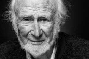 Black-and-white close-up portrait of an elderly man with a long white beard, shaggy white hair and a deeply lined face, gentle quiet gaze.