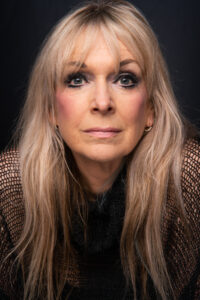 Black-and-white close-up portrait of an older blonde woman with long hair, a dark mesh top and a direct calm gaze, against a dark background.