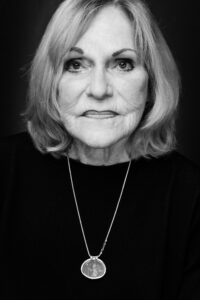 Black-and-white close-up portrait of an older blonde woman with shoulder-length hair, a dark sweater and an ornate medallion pendant on a fine silver chain.