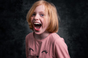 Colour studio portrait of a young girl with ginger hair laughing broadly, wearing a pink T-shirt with a cat-face design against a textured grey backdrop.