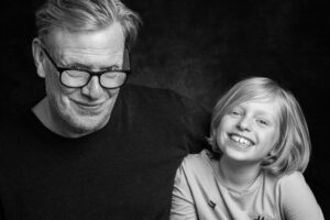Black-and-white studio portrait of an older man with glasses and a young blonde girl close together, both smiling broadly at the camera.