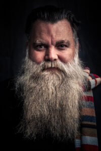 Studio portrait of an older man with a long flowing white beard, direct gaze and a colourful striped scarf, against a dark backdrop.