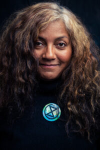 Warm studio portrait of a smiling woman with long curly hair, wearing a black top with an Extinction Rebellion enamel pin.
