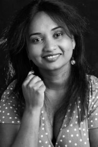 Black-and-white studio portrait of a smiling woman with long dark hair, a polka-dot dress and drop earrings, against a dark background.