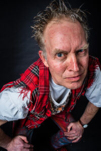 Studio portrait of a man with spiky red-orange hair, a white T-shirt and red tartan plaid wrap, leaning forward with an intense gaze.