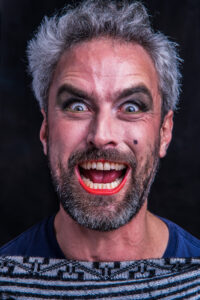 Theatrical portrait of a bearded man with dark eye makeup and red-painted teeth, mouth open mid-shout, against a dark studio backdrop.