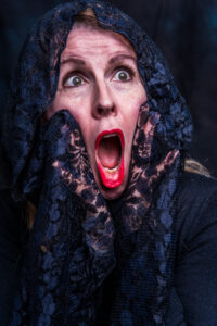 Theatrical studio portrait of a woman in a black lace shawl over head and hands, mouth open in a dramatic scream with red lipstick.