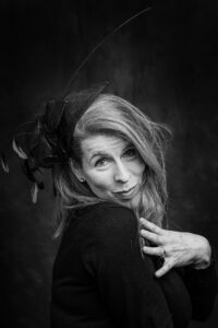 Elegant black-and-white studio portrait of a woman with flowing hair, a black quill-feathered fascinator and a black sweater, glancing playfully.