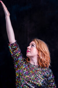 Studio portrait of a woman in a multicoloured sequin top with strawberry-blonde hair, arm raised dynamically against a dark background.