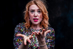 Character portrait of a woman in a multicoloured sequin top with strawberry-blonde hair, hands reaching toward camera with an intense expression.