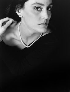 Classical black-and-white studio portrait of a woman with long dark hair, a pearl necklace, and a soft thoughtful gaze against a dark background.