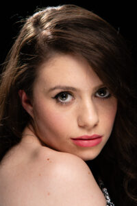 Colour studio portrait of a young woman with wavy brown hair glancing back over her shoulder, soft pink lipstick and a fine chain necklace.