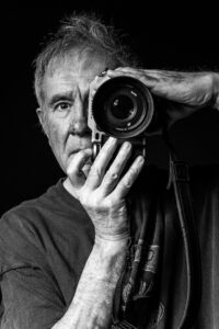 Black-and-white portrait of a photographer holding a medium-format camera to his face, one eye looking past the lens.