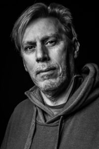 Black-and-white studio portrait of a man with swept-back hair, an eyebrow piercing and a grey hoodie, with a quiet contemplative expression.