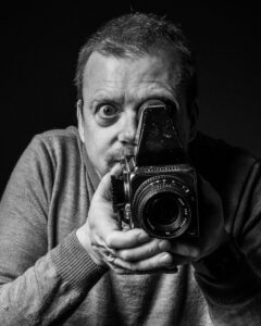 Black-and-white self-portrait of the photographer, wide-eyed behind a vintage medium-format camera held at face level.