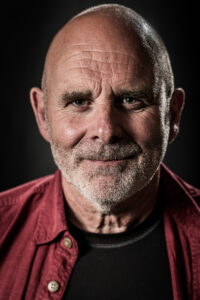 Colour studio portrait of a bald older man with grey stubble and a warm smile, wearing a red shirt over a dark T-shirt.