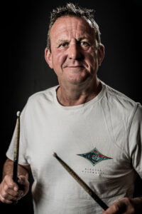Studio portrait of a drummer with grey spiky hair, holding drumsticks and wearing a white T-shirt.
