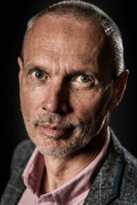 Colour studio portrait of a man with short grey hair, light stubble, wearing a pink shirt under a grey tweed jacket, looking directly at camera.