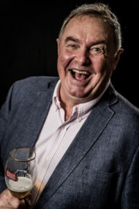 Colour studio portrait of a man laughing heartily while holding a beer glass, wearing a grey blazer over a pink shirt.