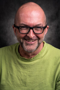 Colour studio portrait of a bald man with grey stubble, black-rimmed glasses and a lime green T-shirt, smiling broadly.
