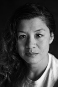 Black-and-white studio portrait of a woman with shoulder-length curly hair, freckles and a calm direct gaze, wearing a white shirt.