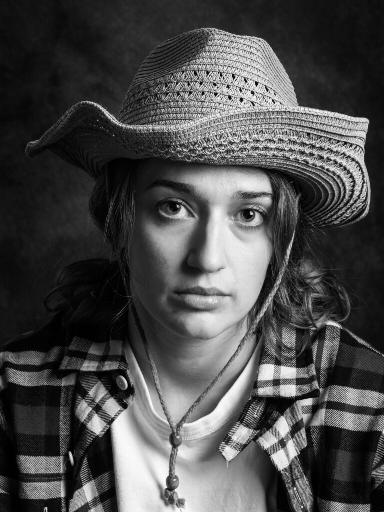 Black-and-white studio portrait of a young woman in a straw cowboy hat and plaid shirt, with a wooden pendant and serious expression.