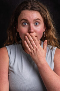 Colour studio portrait of a young woman with curly auburn hair and wide blue eyes, hand covering her mouth in a startled gesture.