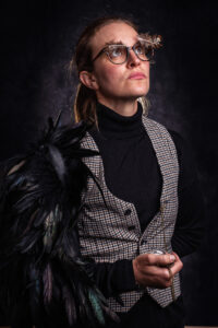 Studio portrait of a woman with glasses adorned with a feather quill, a tweed waistcoat and dark feathered cape, holding a pocket watch.