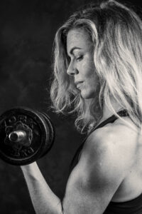 Black-and-white studio portrait of a blonde woman in profile lifting a dumbbell, focused expression, against a dark studio background.