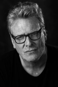Black-and-white studio portrait of an older man with spiky grey hair and black-rimmed glasses, looking down with a thoughtful expression.