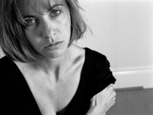 Black-and-white studio portrait of a woman with a fair bob hairstyle, scoop-neck black top, and an intense direct gaze in natural light.