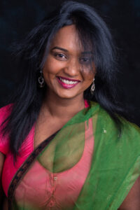 Colour studio portrait of a smiling woman wearing a pink and green sari with chandelier earrings, against a dark background.