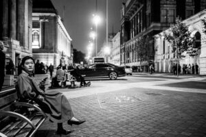 Black-and-white street scene of a woman sitting on a bench at night looking at her phone, the stone classical buildings of Exhibition Road behind, South Kensington.