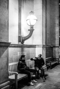 Black-and-white street scene of two men sitting on a bench under an ornate cast-iron wall lamp at night, against the stone columns of Imperial College London.
