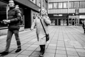 Black-and-white candid of two pedestrians passing on a Stevenage high street, a William Hill betting shop and a Rosies storefront in the background.