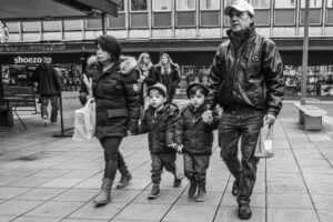Black-and-white candid of an older man holding two small boys' hands, a woman alongside, walking past Stevenage high-street shops.