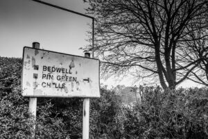 Black-and-white photograph of a weathered painted road sign reading "Bedwell · Pin Green · Chells" beside a bare tree on a country lane.